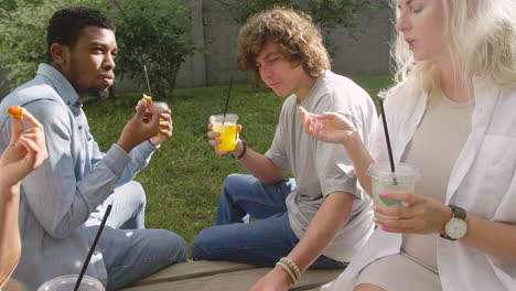 group of multiethnic friends spending time together, eating delicious nuggets and drinking refreshing juice, sitting in a wooden bench in a park and talking to each other