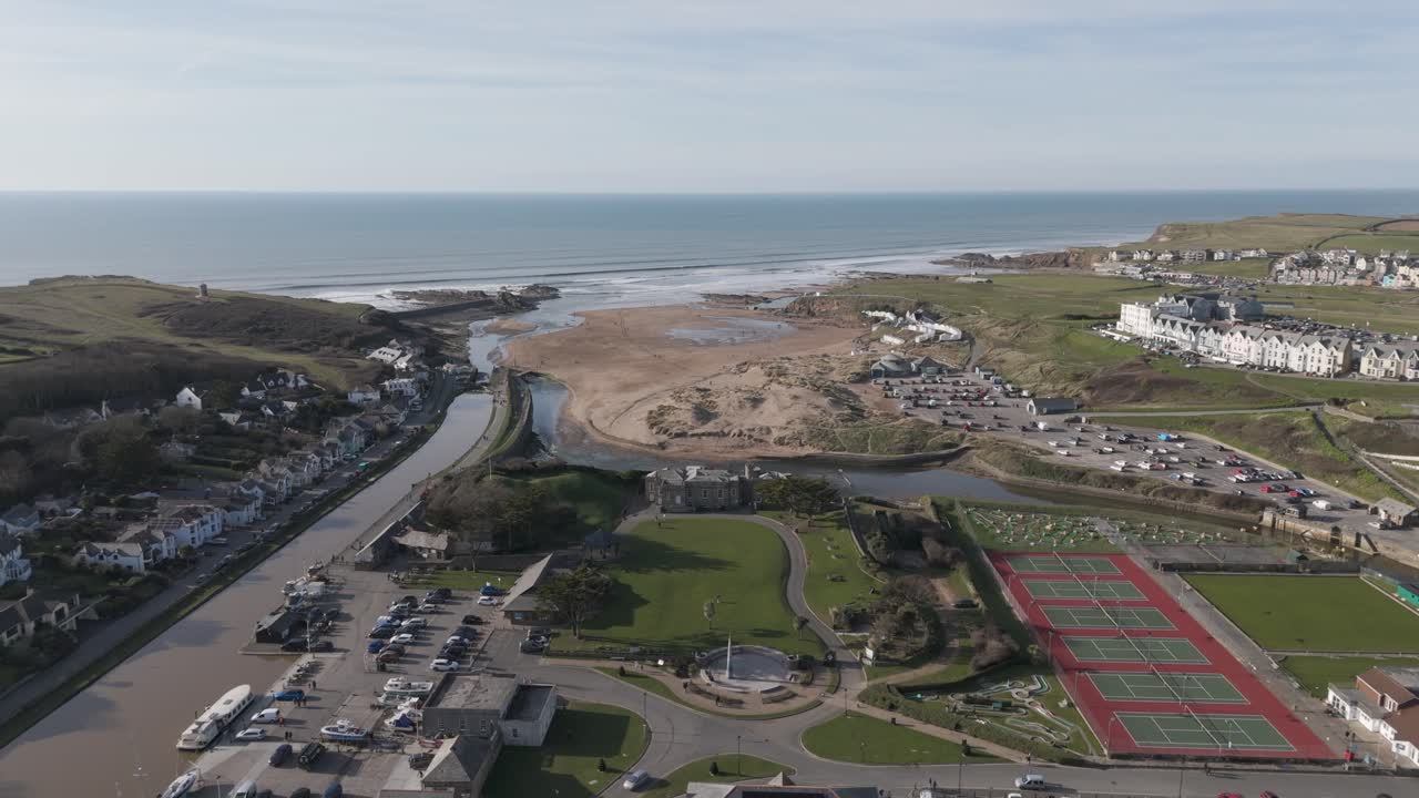 High aerial over Bude coastal town where sandy beach, recreation fields and holiday parks meet Atlantic surf between rocky headlands on Cornwall’s north coast