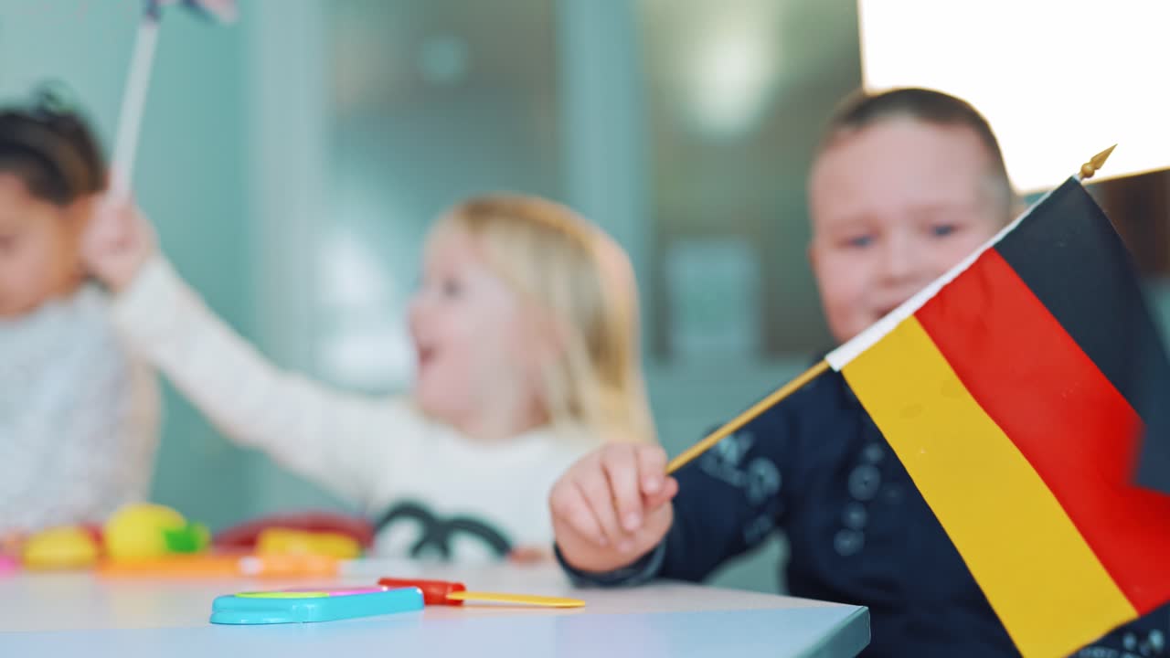 Little boy and a girl with flags at class. German and British flags in the hands of small children sitting at the table. Kids wave flags indoors.