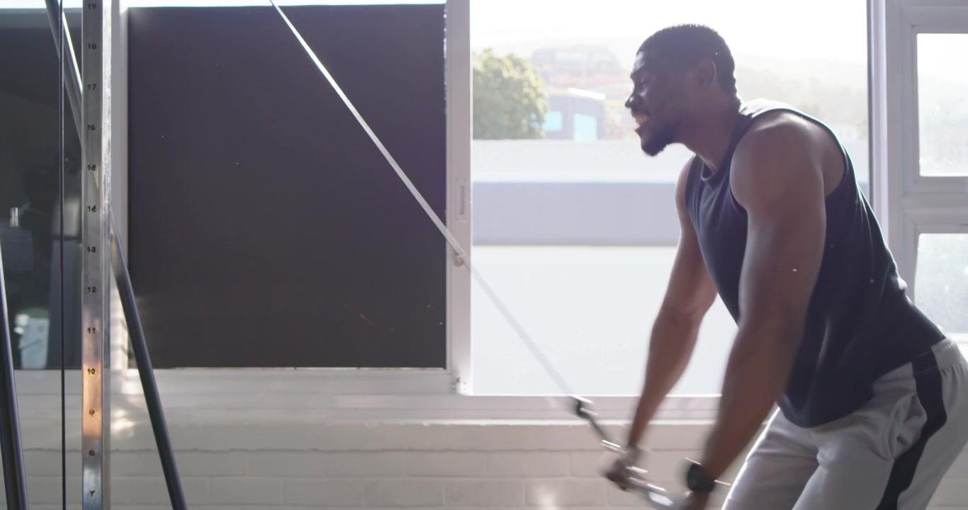 African man at gym in tank grabbing cable handle, leaning forward, pulling toward hips for fitness