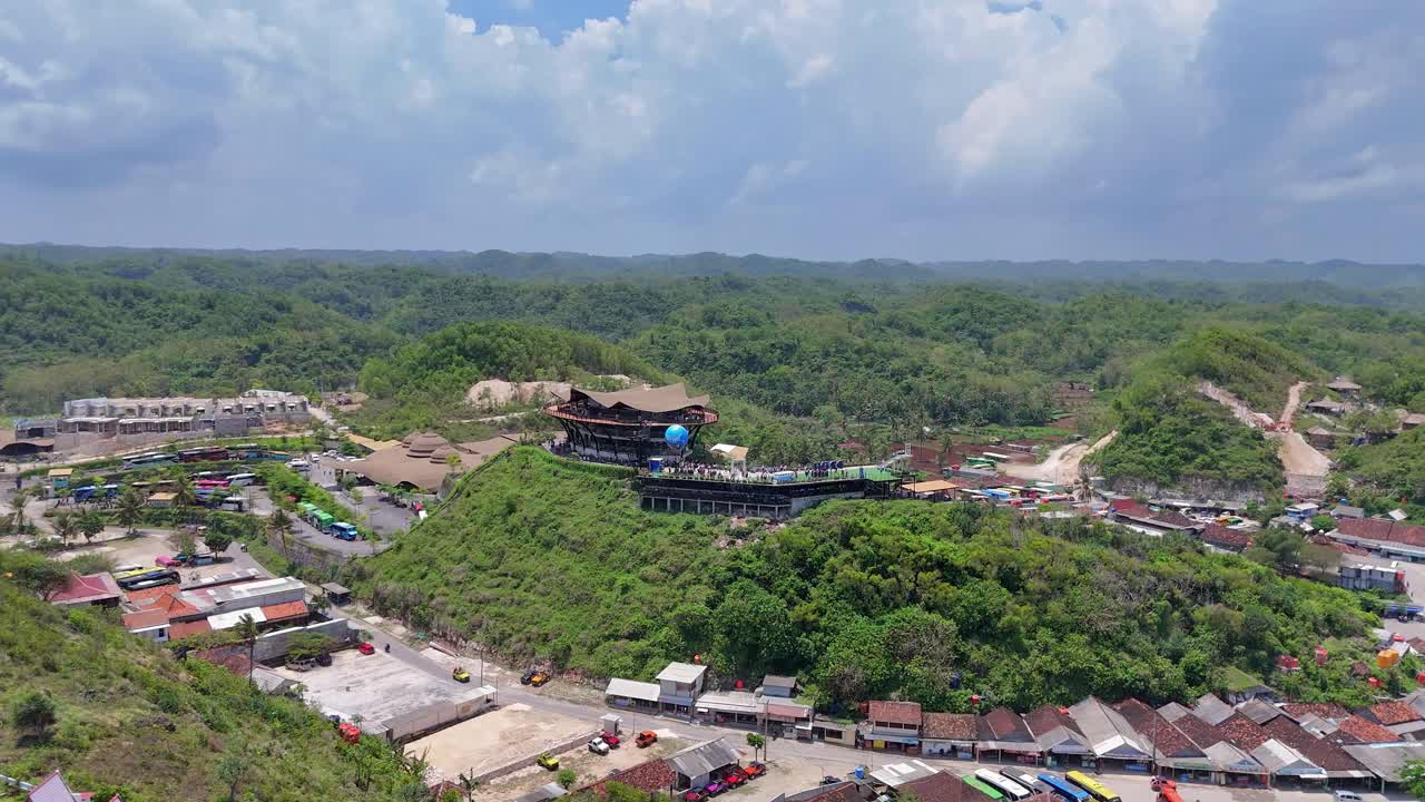 Drone aerial view of Drini Park on Drini Beach, Yogyakarta, Indonesia, showcasing a scenic hilltop building surrounded by lush green hills and coastal landscape under a bright blue sky