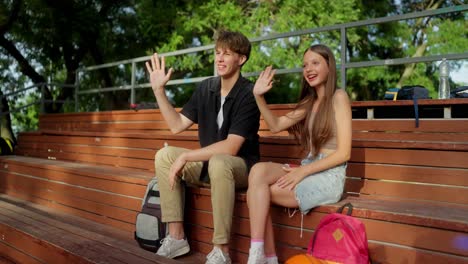 two teenagers sitting on a bench in a park and waving