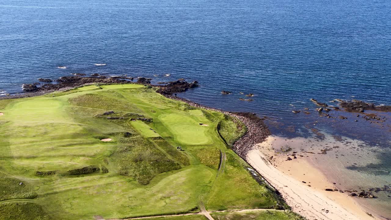 Drone glides above green golf course, sandy beach, and rocky coastline in bright daylight