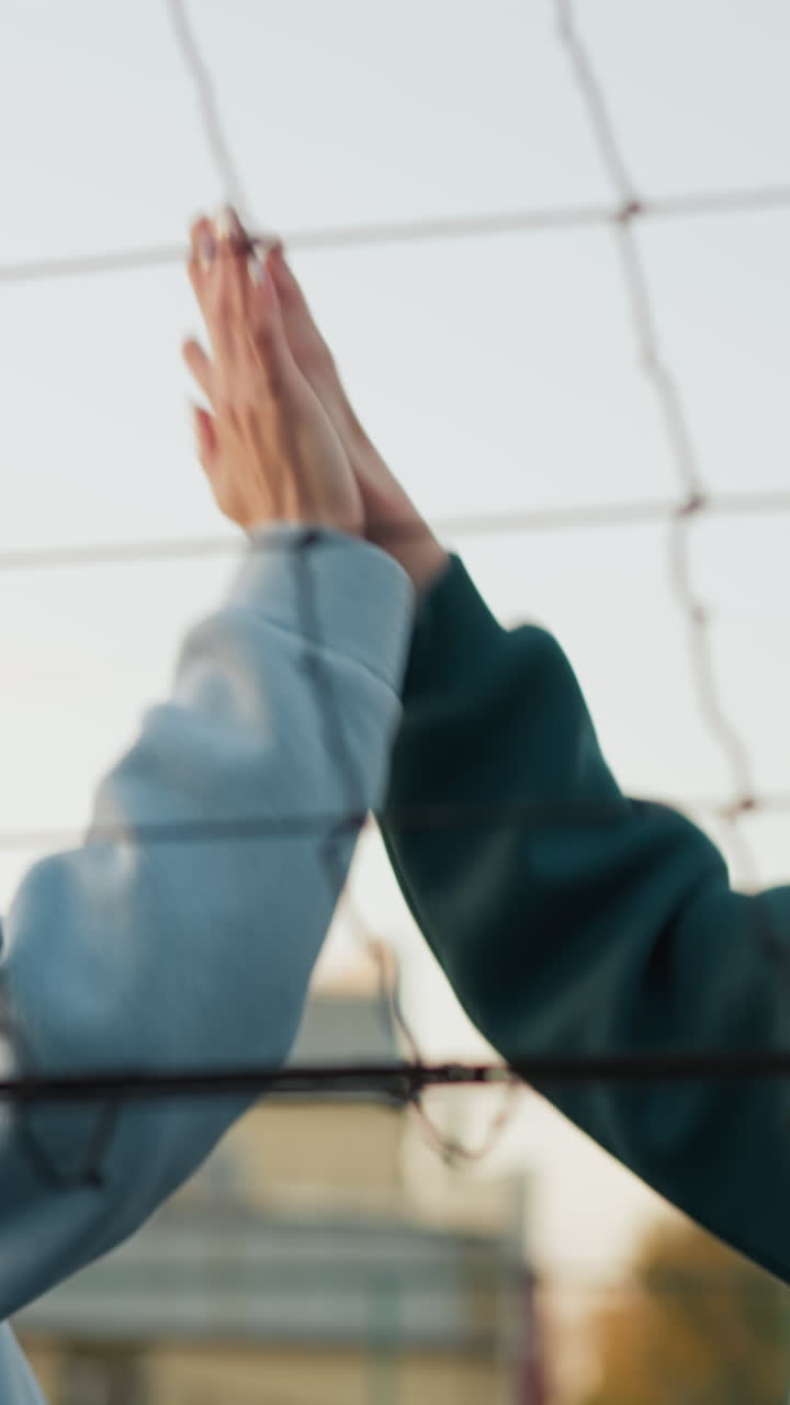 primer plano de compañeros de equipo con sudaderas verdes y azules saltando juntos, enviando la pelota de voleibol de vuelta a la red con alegría mientras se unen de manos, edificios visibles en el fondo durante el partido al aire libre