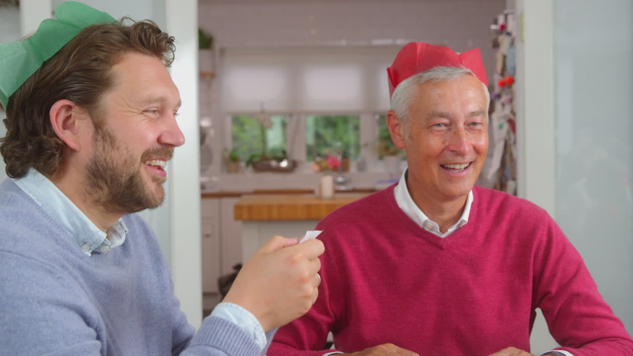 Padre anziano con figlio adulto che leggono barzellette dai Christmas cracker mentre mangiano insieme