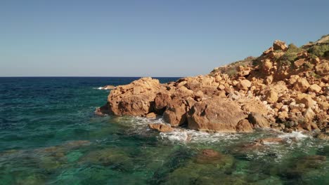 aerial forward moving shot of a seashore with beautiful turquoise water of the mediterranean sea splashing on the red coloured rocks