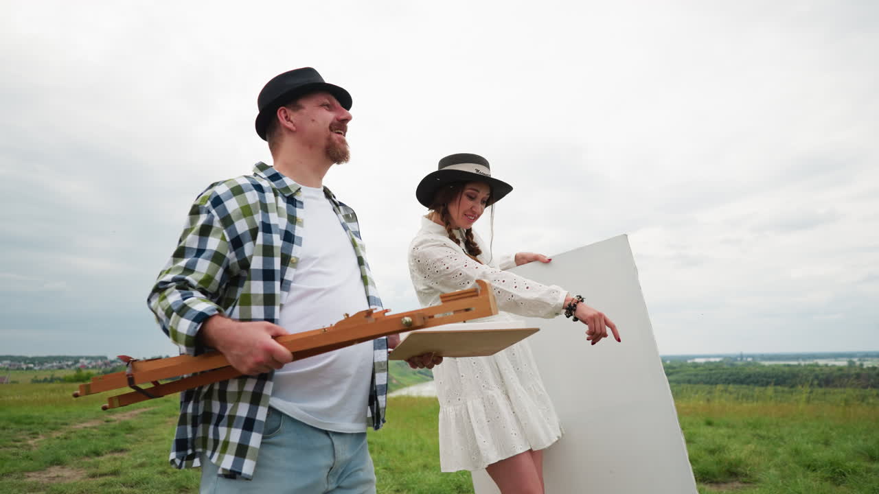 мужчина и женщина в повседневной художественной одежде ходят по травяному полю с художественными принадлежностями. мужчина носит клетчатую рубашку, джинсы и черную федору, держа в руках шезлонг. женщина, улыбаясь,