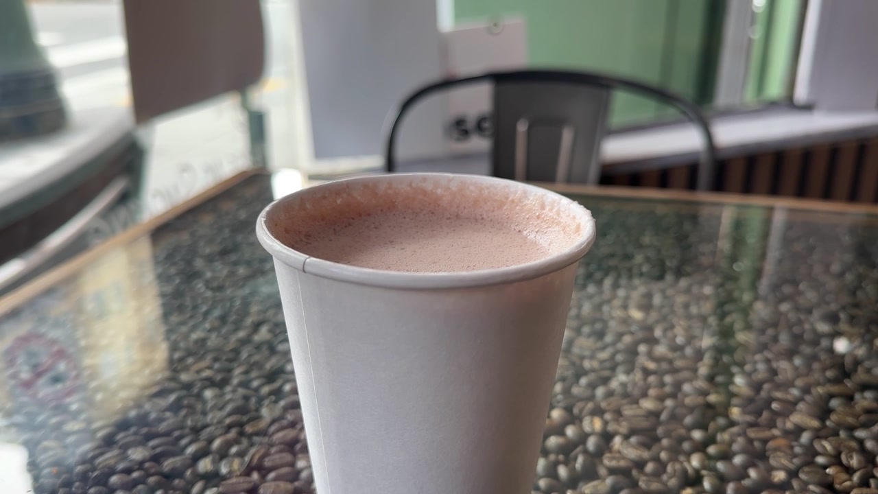 Deliziosa tazza di cioccolata calda fumante in una tazza di carta bianca appoggiata su un tavolo unico pieno di chicchi di caffè in un accogliente caffè a San Francisco, con una sedia e una finestra sullo sfondo.
