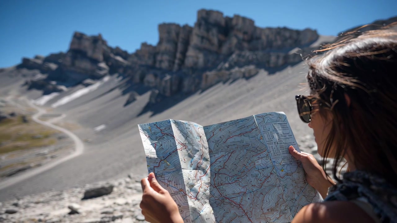 Aufnahme einer Reisenden, die eine gefaltete topografische Karte hält, während sie in einem zerklüfteten Alpental mit felsigen Gipfeln und einem Feldweg unter klarem blauem Himmel wandert.