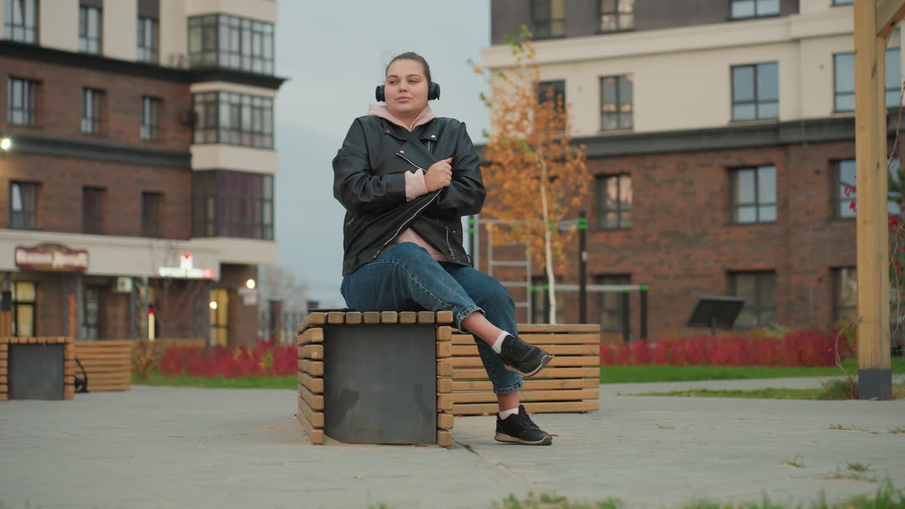 Woman seated on urban bench outdoors adjusts jacket to stay warm while wearing headphones and listening to music in cool weather, surrounded by autumn colors