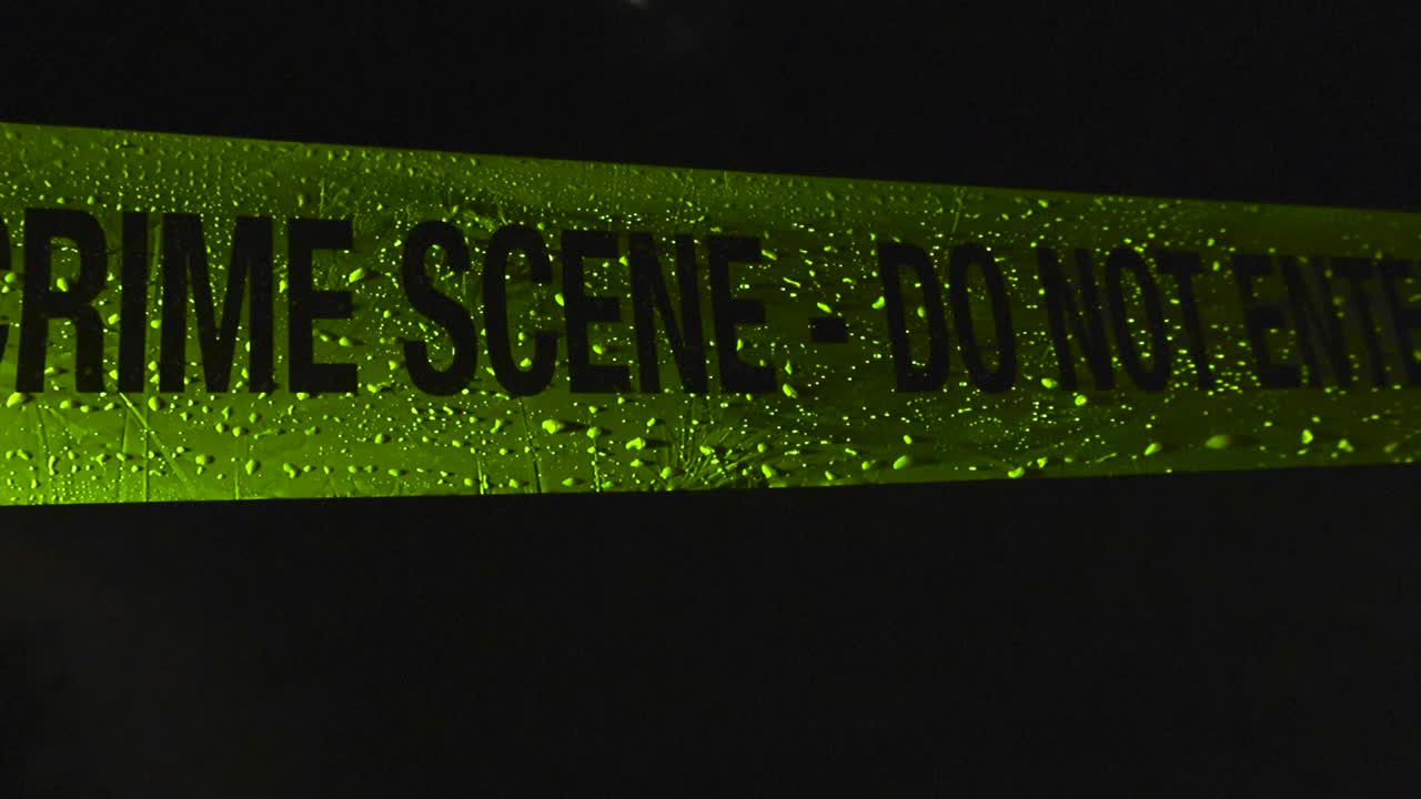 Closeup or close up view of a yellow vibrant colored Crime Scene Do Not Enter police caution tape ribbon moving in the wind while being covered by raindrops in a dark black night, light on ribbon