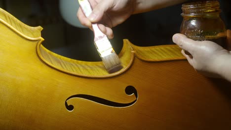 skilled luthier carefully applying self prepared varnish to wooden cello ribs, using traditional brush and craftsmanship in sunlit workshop setting, real time