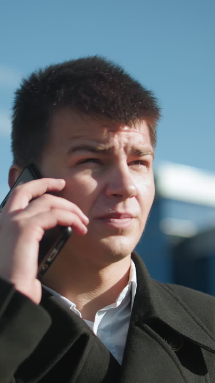 검은 코트를 입은 계약자는 야외에서 전화 대화를 나누고, 의사소통하는 동안 눈에 띄게 머리를 흔들며, 현대적인 유리 건물과 함께 도시 환경에 서 있습니다.