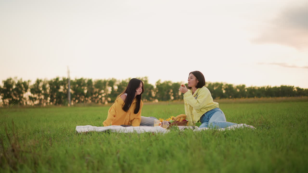 해 질 녘 들판에서 피크닉을 즐기는 두 여성이 이야기를 나누고 있어요. 대학 친구인 그들은 담요 위에서 과일을 나눠 먹으며 부드럽게 웃고 있고, 편안한 자세로 넓은 풀밭에 앉아 있어요. 시골 지평선 위로 부드러운 황금빛 햇살이 비치고, 캐주얼한 데님을 입고 있네요.