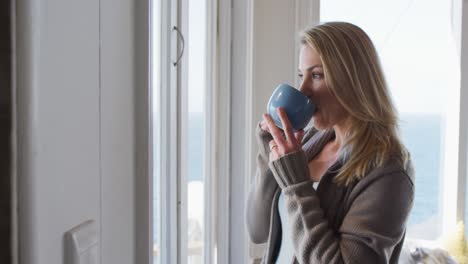 mujer caucásica madura mirando por la ventana y bebiendo café