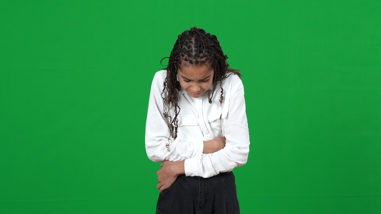 Upset teen African American girl suffering stomach ache on green screen. Portrait of sad unwell teenager posing at chromakey background. Medical problems on periods or food poisoning.