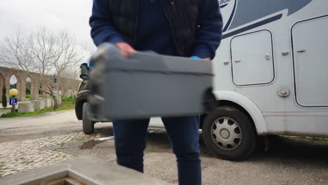 a man shakes a plastic toilet container in front of a caravan