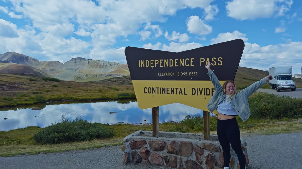 Smiling woman poses for a photo at the Continental Divide sign at Independence Pass in Colorado, with Rocky Mountain scenery in the background on a summer day
