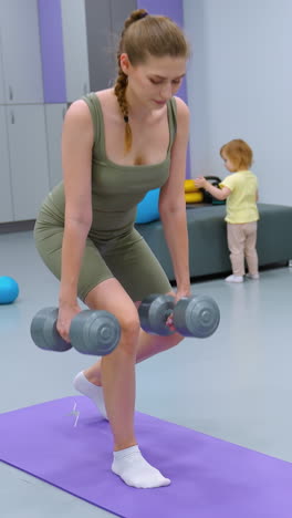 mujer enfocada realiza entrenamiento de fuerza con pesas en una alfombra de yoga mientras el niño pequeño juega en el fondo, equipos de fitness, alfombras de ejercicio y niño lúdico crean un entorno de gimnasio dinámico