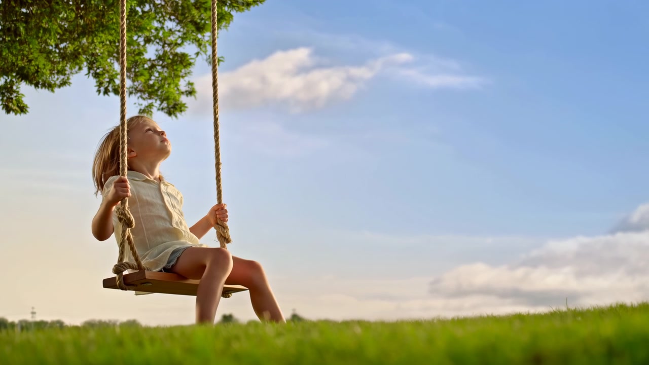 Un bambino siede su un'altalena di corda in un parco verde sotto un albero frondoso, visto da un'angolazione bassa con erba in primo piano e un cielo azzurro brillante con nuvole sullo sfondo.