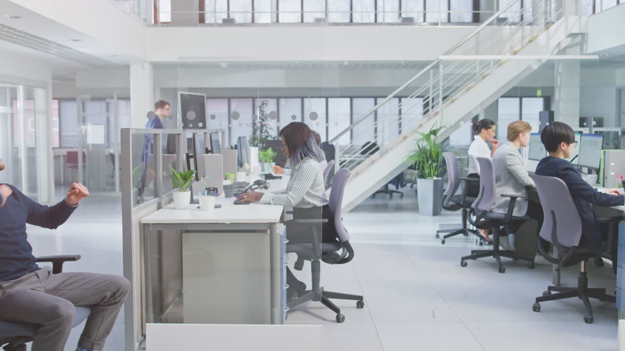 쾌활한 직원은 그의 잘생긴 동료를 현대 사무실 공간에서 데스크탑 컴퓨터에서 일하는 다양한 비즈니스 사람들과 함께 책상 줄 에 있는 의자에 밀어 넣습니다.