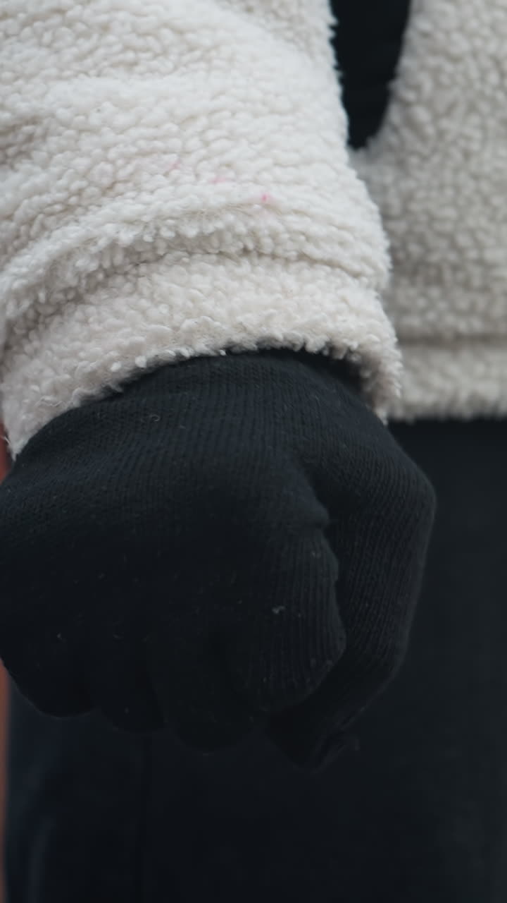 검은 장갑, 겨울 재, 검은 바지를 입은 사람이 손목을 회전시키고, 눈이 흐릿한 배경과 불분명한 인물들을 보여주는 클로즈업 측면 시각