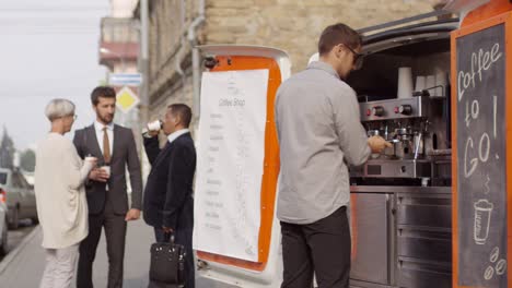 male barista working on coffee machine in mobile van, and group of multiethnic office workers in business suits standing in city street, drinking from disposable cups and talking