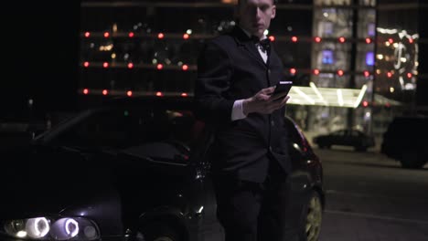 hombre de negocios en traje usando un teléfono inteligente al lado de un lujoso coche ejecutivo y una gran ventana en el estacionamiento. luces en el fondo