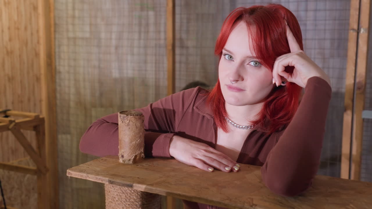 Donna dai capelli rossi appoggiata al tiragraffi in un laboratorio di bamboo, con le braccia appoggiate ai pali in sughero mentre guarda pensierosa la calda luce della finestra: un'artigiana che contempla il design dei mobili