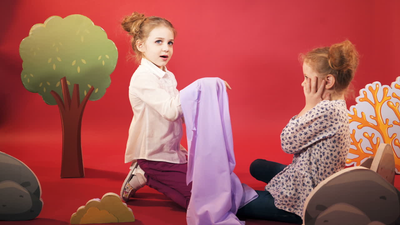 two little sister are playing with a purple fabric on the background of red studio with scenery in the form of trees