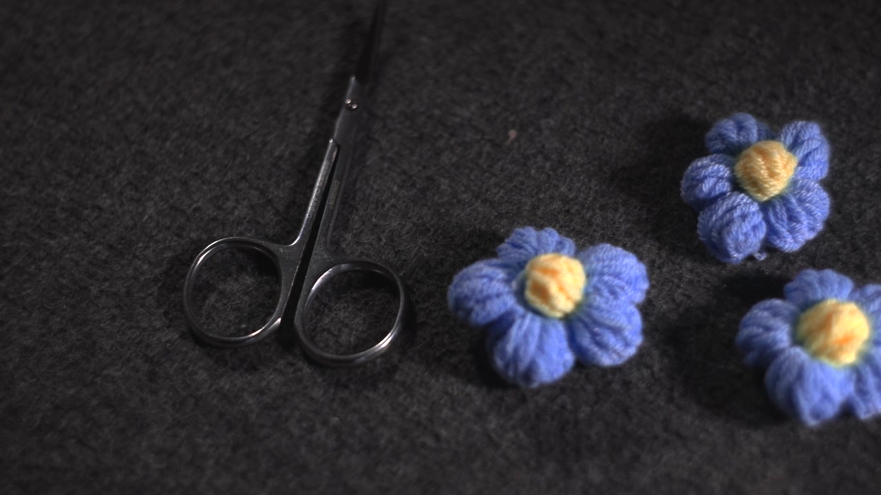 Gros plan d'une main taillant des fleurs en laine avec de petits ciseaux sur un fond de feutre foncé, pétales bleus crochetés