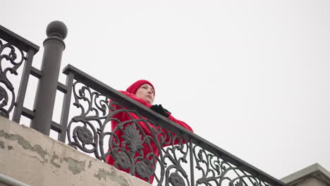 mujer con capucha roja y gorra descansa en una barandilla de hierro decorativa bajo un cielo brillante, mostrando un intrincado diseño de valla y un ambiente invernal urbano sereno