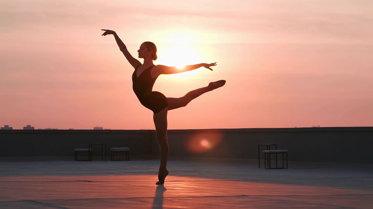Ballerina in body scuro esegue un'arabesque in equilibrio su un tetto al tramonto, bilanciata su una punta con la gamba posteriore estesa, braccia sollevate, cielo luminoso, riflessi tenui e un paesaggio urbano calmo