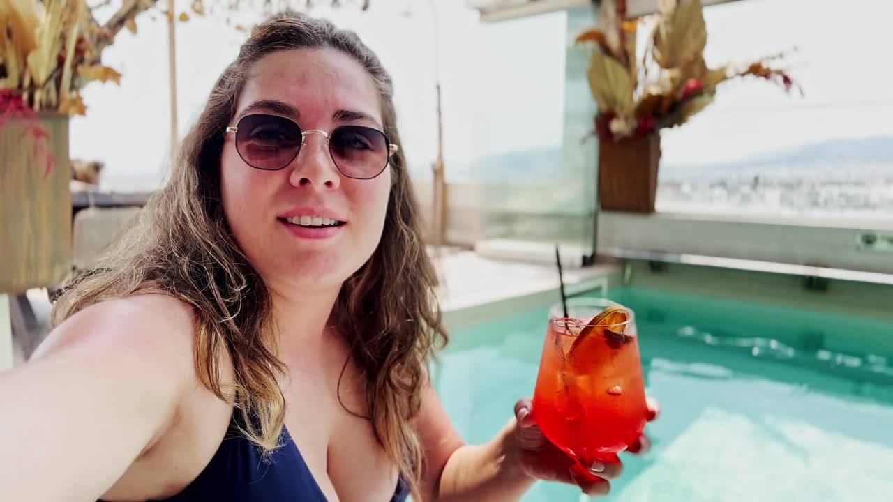 A Happy Young Woman with a Cocktail Talking to the Camera at a Poolside Bar