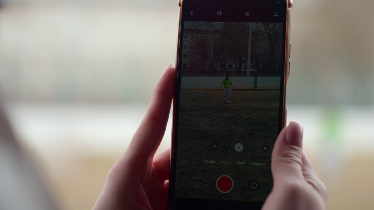 primer plano de un niño grabando con un teléfono en un campo, con las manos sujetando el dispositivo y el botón rojo de grabar visible, fondo de ventana borroso, luz interior suave, agarre firme capturando movimiento y recuerdos distantes para más tarde