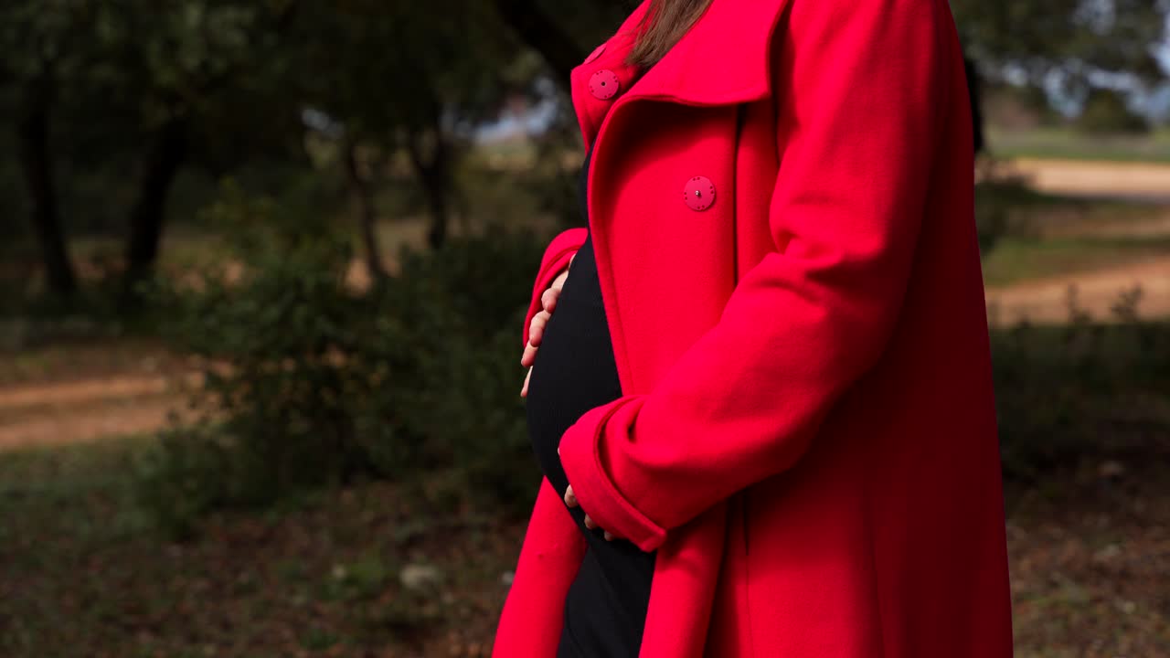 Pregnant woman stands in forest path while unbuttoning red coat to reveal belly