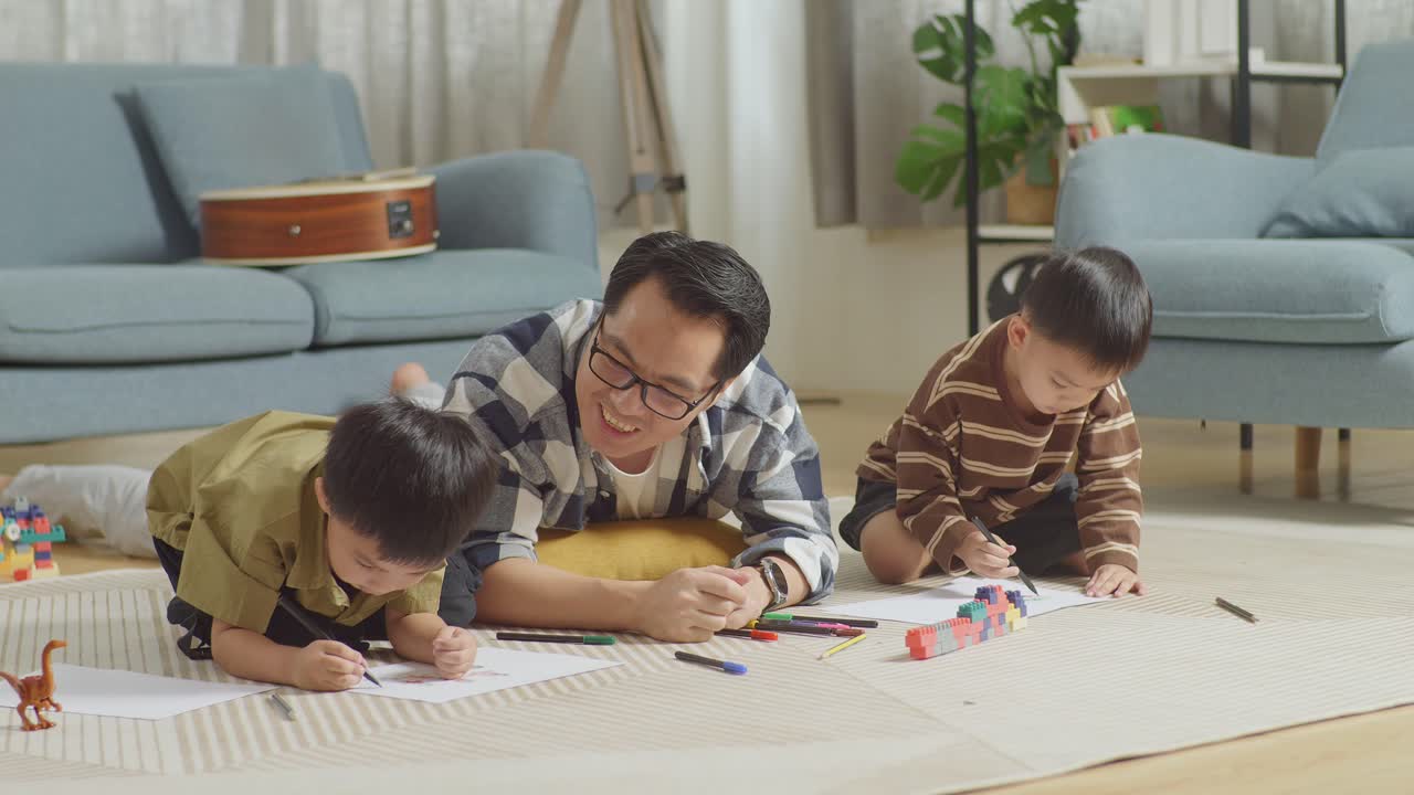 아시안 아버지와 아들의 몸이 가득 차있는 방의 바닥에 플라스틱 장난감 벽돌이 집에서 함께 그림을 그리고 있습니다. 웃는 아버지는 그림을 연습하는 아들과 얘기하고 있습니다.
