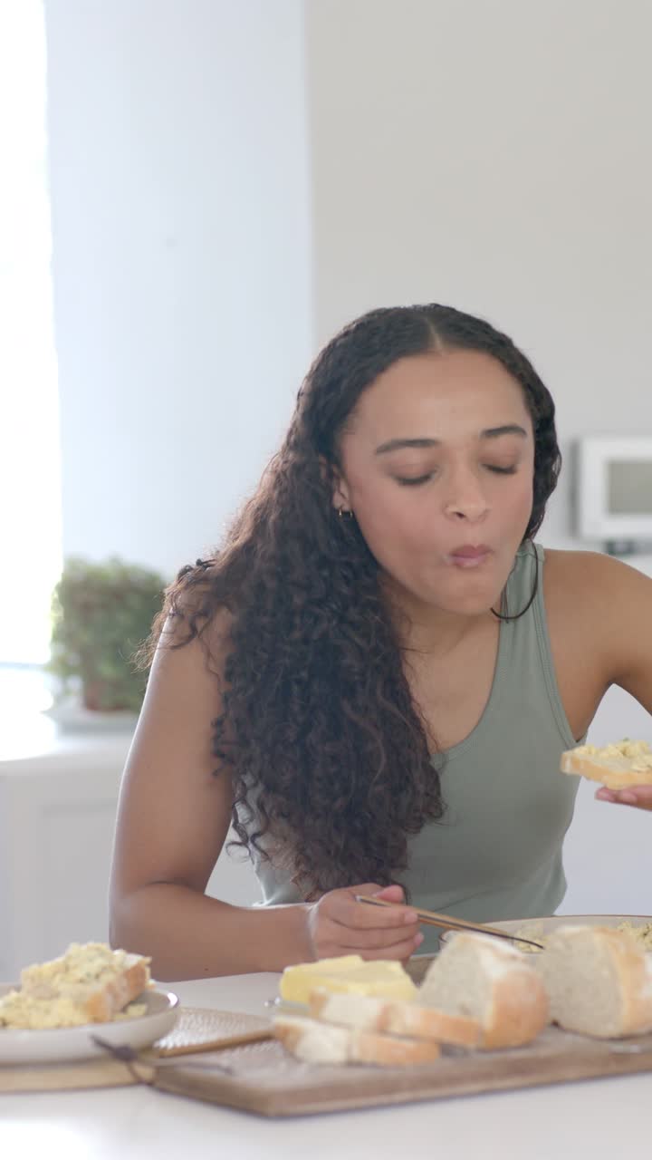 Mujer afrodescendiente con camiseta de tirantes untando paté con un tenedor en el pan en la encimera de la cocina, probando.