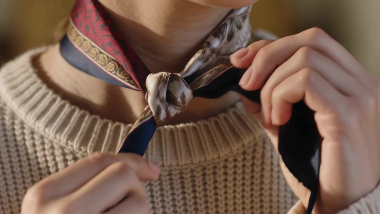 Una donna annoda una sciarpa di seta al collo, sopra un maglione a coste color crema, a casa, per completare il suo stile.