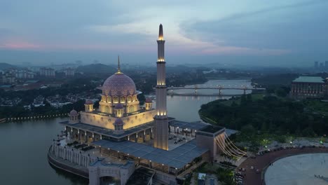 stunning aerial view of the illuminated putra mosque in putrajaya, malaysia, surrounded by serene waters and cityscape during dusk, showcasing its architectural beauty and tranquil ambiance