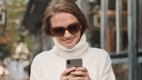 caucasian female in sunglasses using smartphone while walking outdoors.