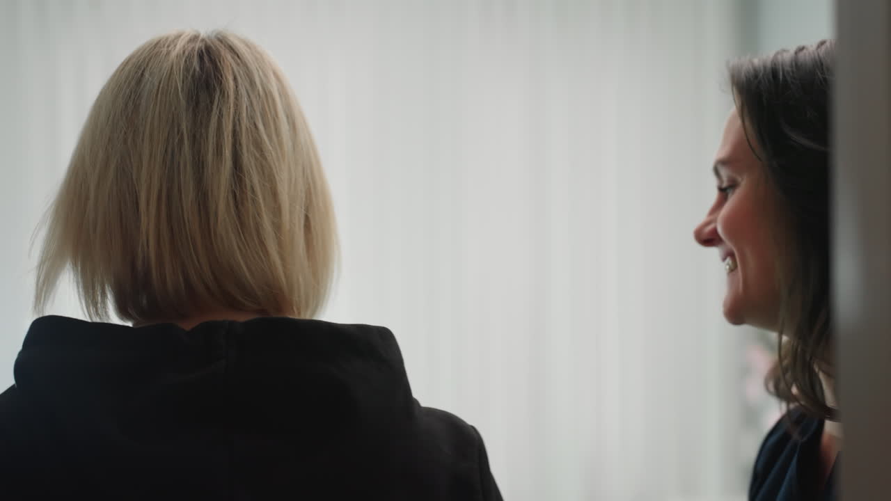 Skin therapist in navy uniform opens door with warm smile to welcome client, then gestures toward cream massage chair in bright white room, creating professional atmosphere before facial treatment