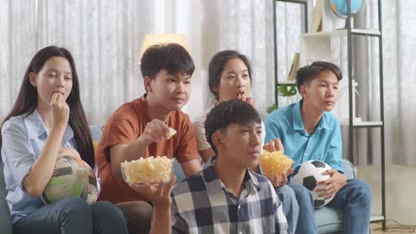 asian teenagers cheering, eating and watching football game on tv at home