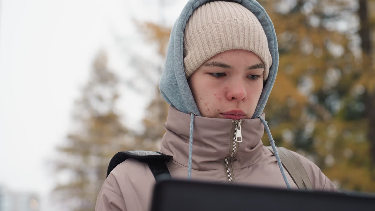 Light-skinned woman in hooded jacket and beanie intently using laptop outdoors in cold weather with blurred background of autumn foliage, showing concentration and calm expression during digital work