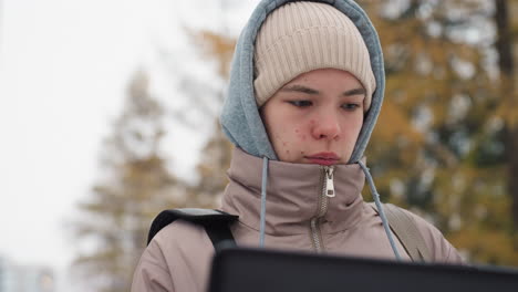 light-skinned woman in hooded jacket and beanie intently using laptop outdoors in cold weather with blurred background of autumn foliage, showing concentration and calm expression during digital work