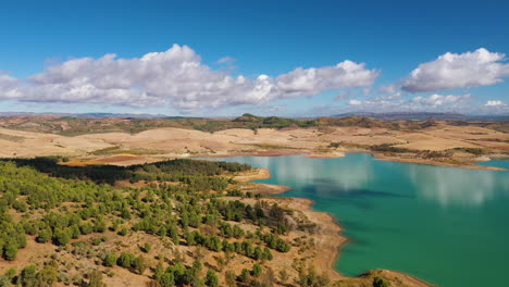 Aerial-drone-shot-of-a-cyan-coloured-lake-surrounded-by-a-rocky-dessert-landscapes-and-forest