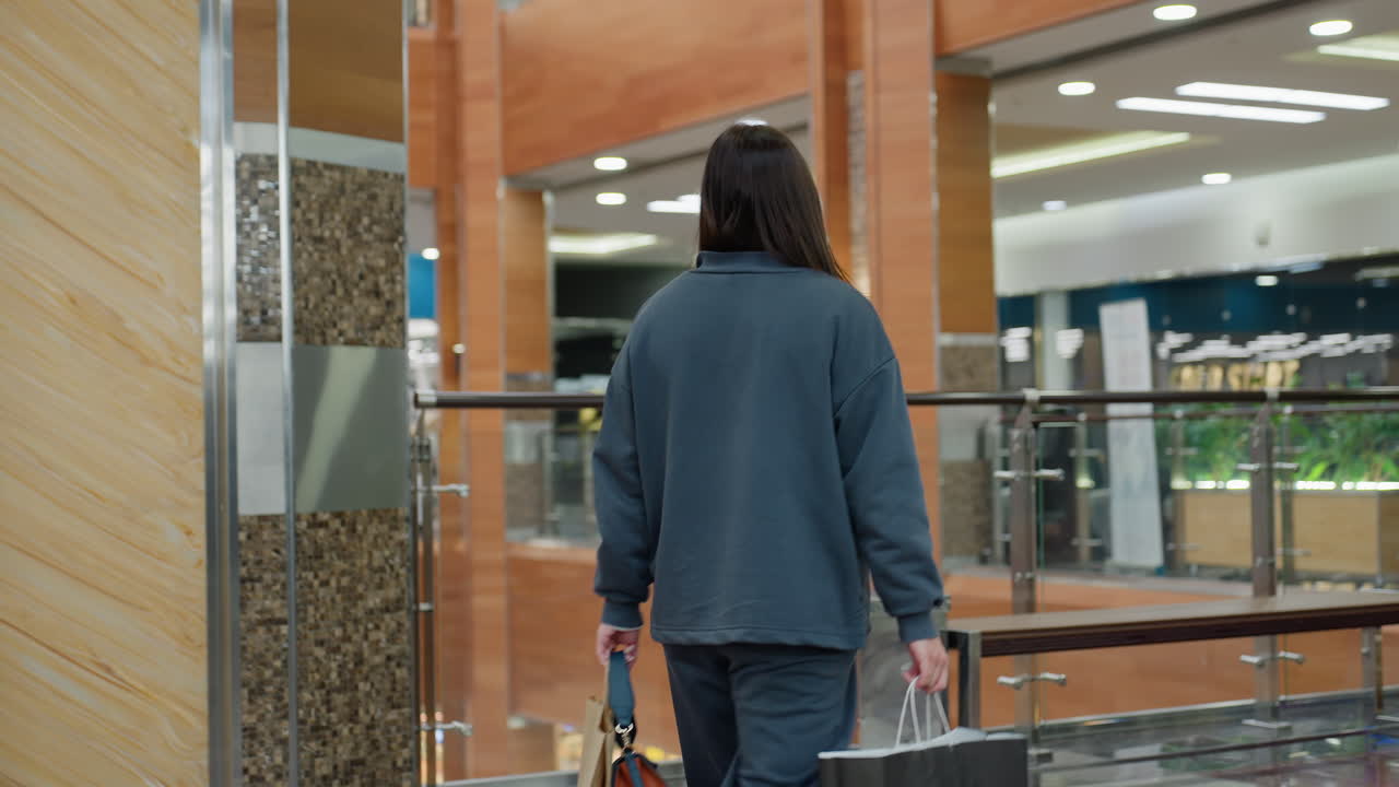 Rear view of shopper walking toward bench inside brightly lit shopping mall, holding black and brown paper bags, preparing to sit in relaxed posture as crowd shops in background behind glass railing