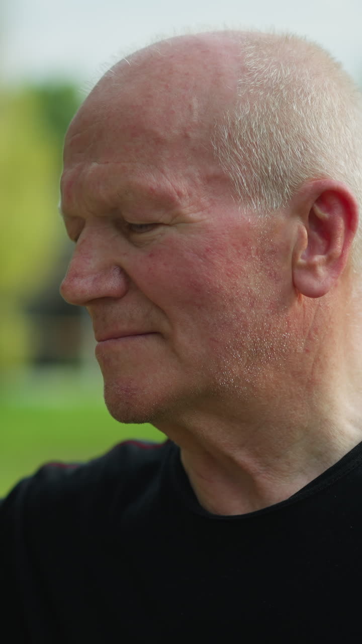 An elderly man in a black top is thoughtfully observing a soccer ball held close to his face, with a blur view of building in the background