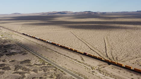 extremely long freight train with hundreds of train cars speeds down a straight train track in the desert