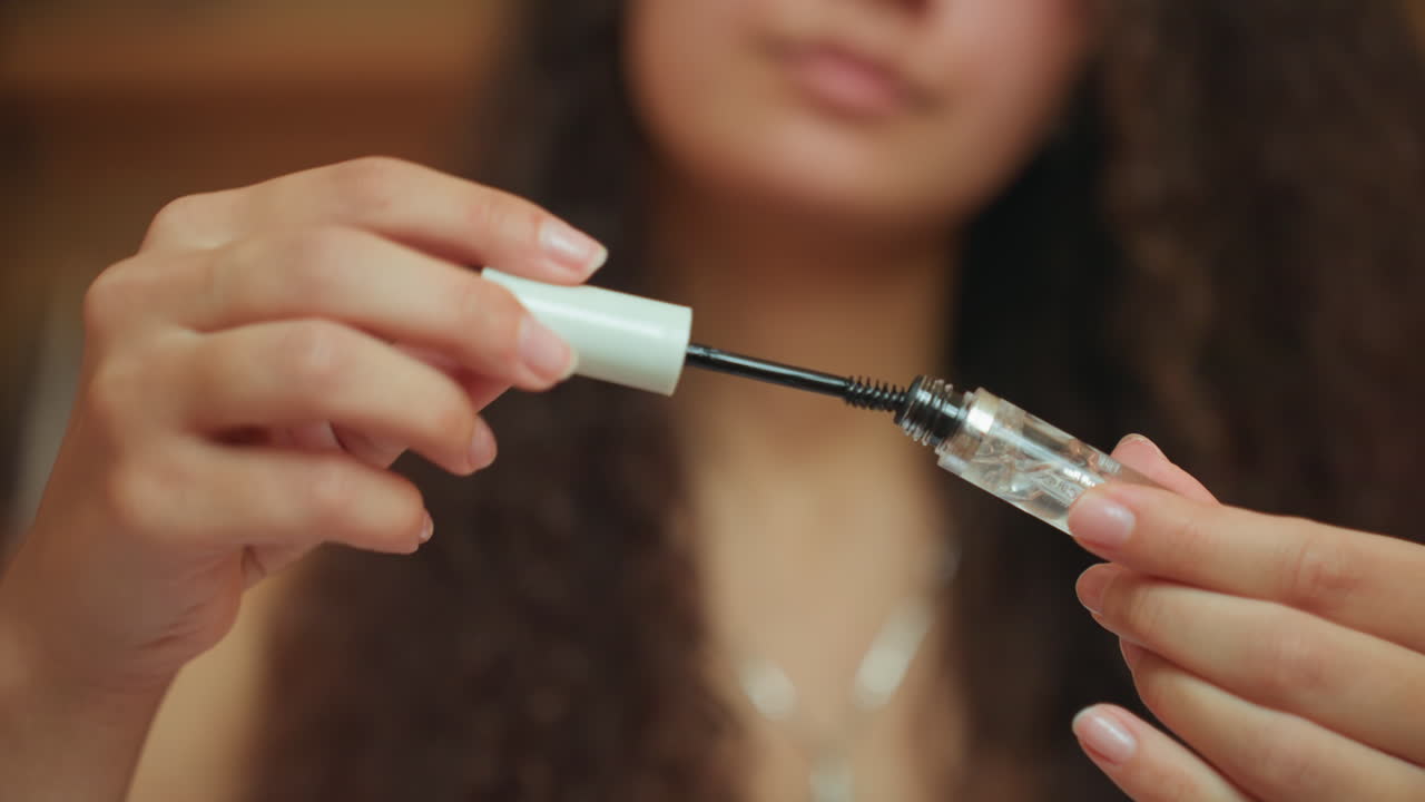 Primer plano de manos aplicando máscara de pestañas en la habitación del hotel, mujer de cabello rizado preparándose para la noche, aplicador sacado de un tubo transparente, pestañas cubiertas con suero transparente, iluminación suave y cálida y fondo difuminado transmiten.